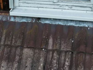 Detailaufnahme eines Daches per Drohne, die einen gerissenen Dachziegel unter einer Fensterbank und verwitterte Metallverkleidungen zeigt.